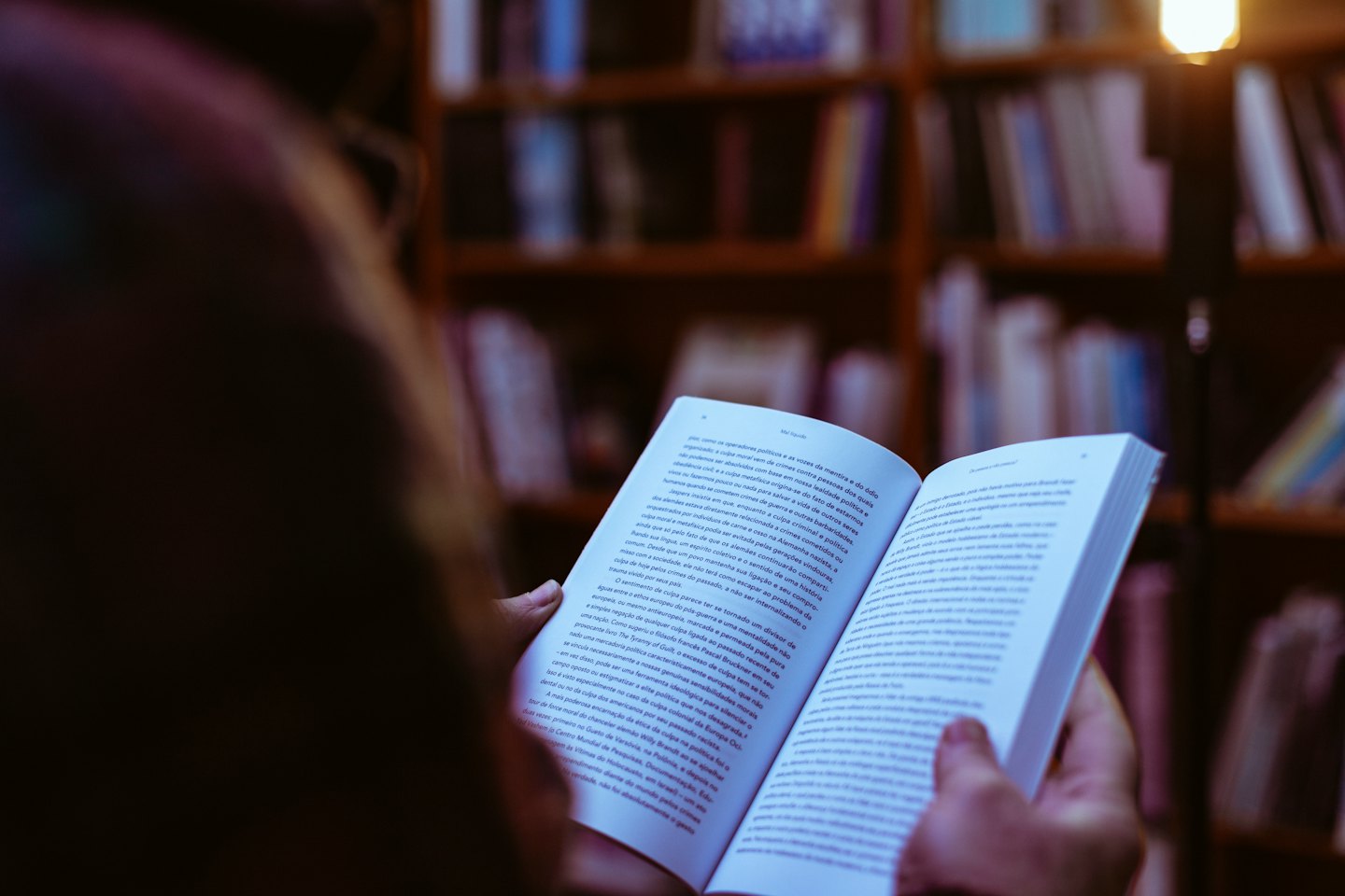 Person liest ein offenes Buch mit unscharfen Bücherregalen im Hintergrund.